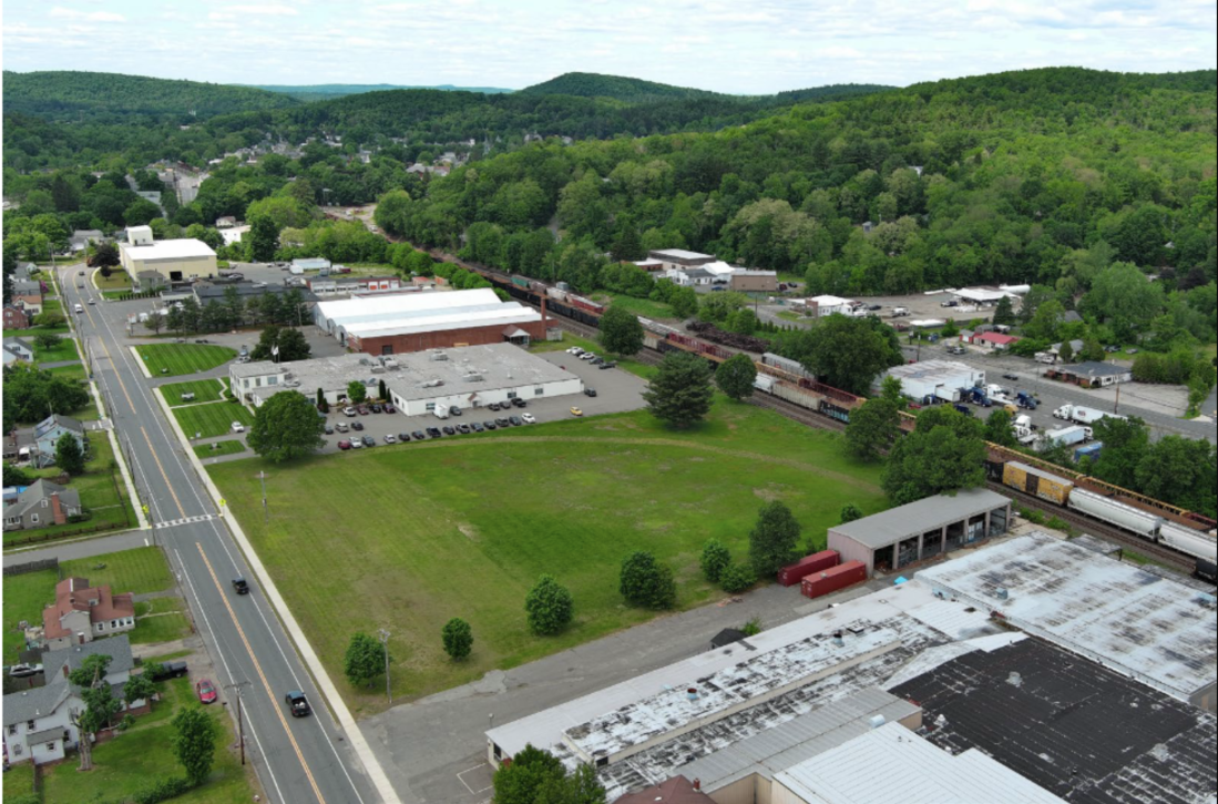 An aerial view of a field between two warehouses, a rail line to the right, and a street with sidewalks in the lower left. In the distance are some green forested hills.