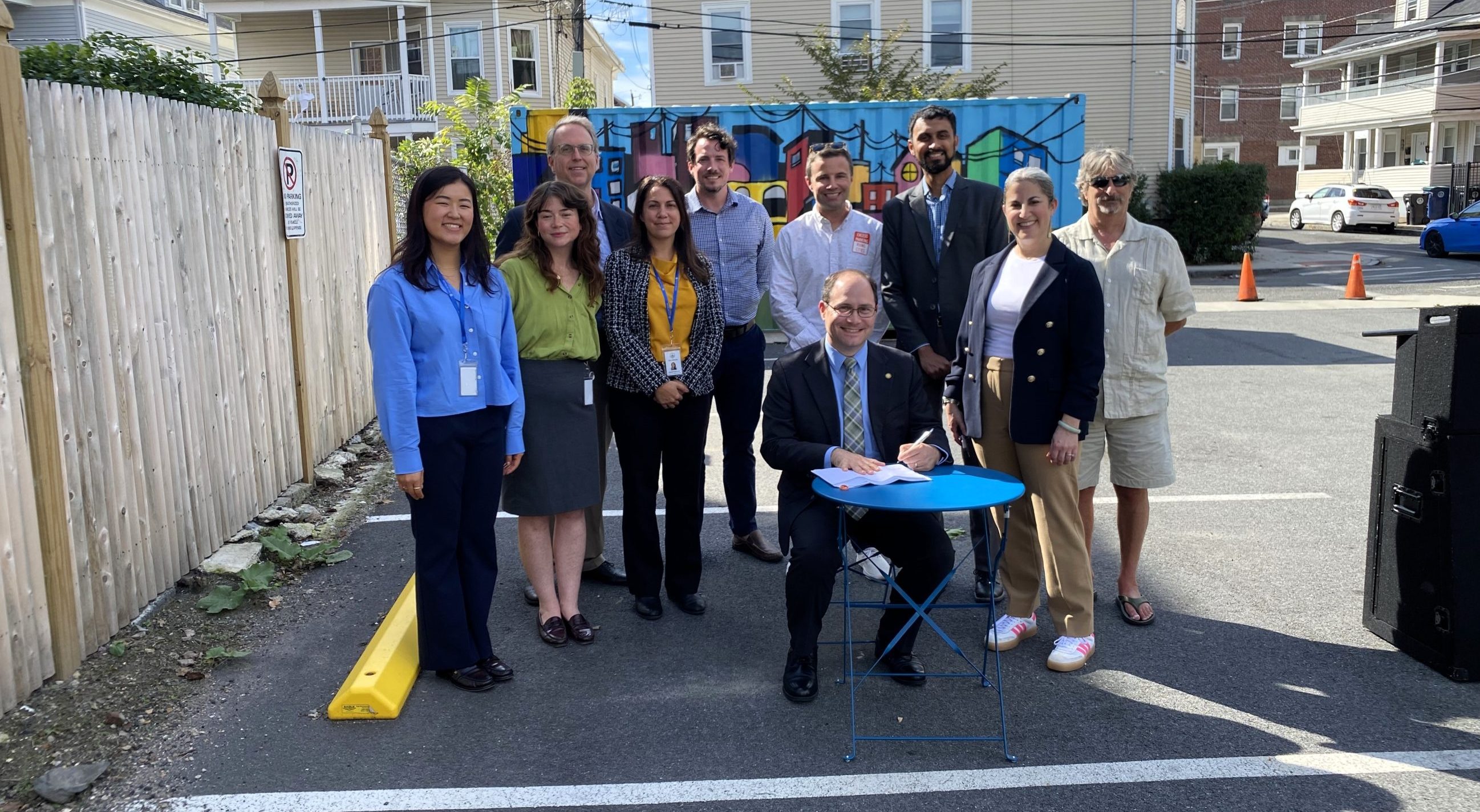 A small crowd of people in business-casual attire surroudn a man in a suit seated at a round table, signing a document. The people are gathered in a parking space in an otherwise empty parking lot surrounded by small apartment houses.