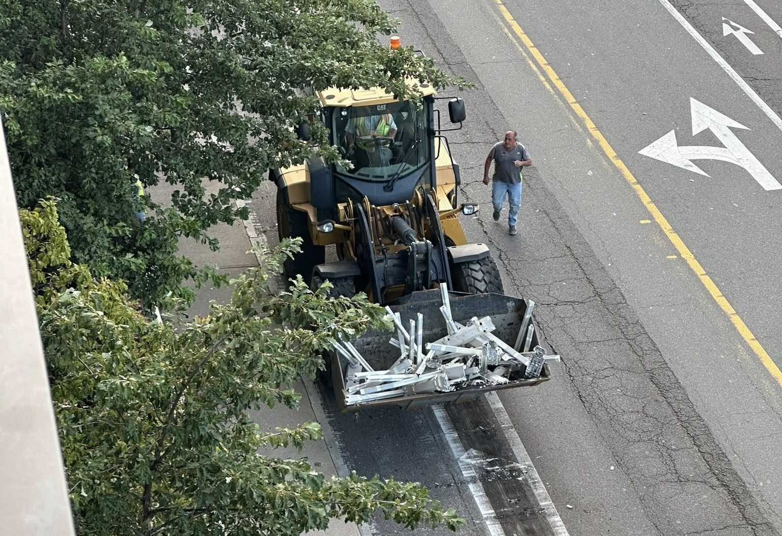 A photo taken from an elevated vantage point of a bulldozer parked in a bike lane with a bucket full of white plastic bollards.