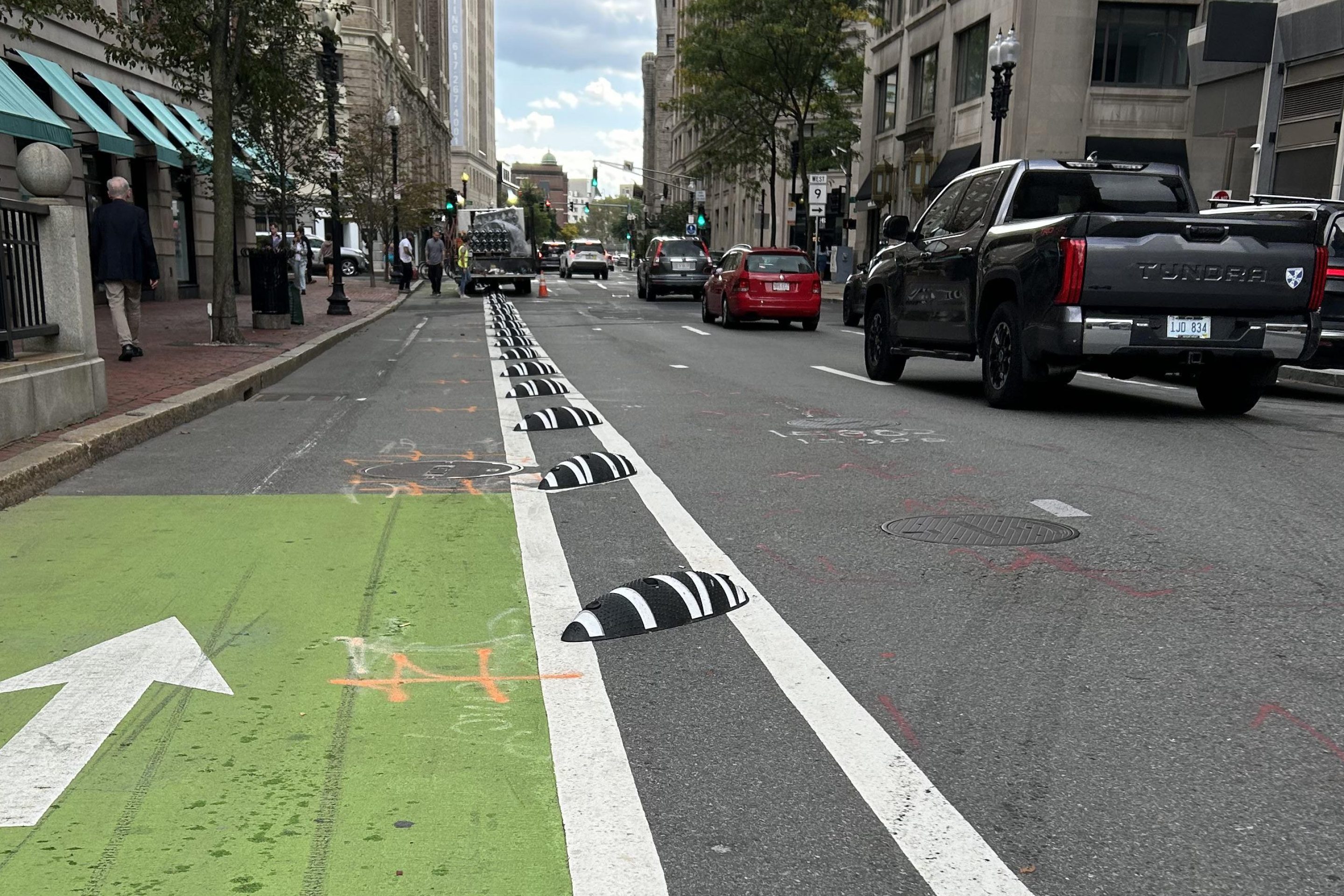 A low-angle view of a city street with a green-painted bike lane to the left, and a row of black-and-white striped plastic mounds installed between two white stripes in the buffer zone between the bike lane and the car lanes to the right. In the distance, workers are installing more of the black-and-white barriers from a truck parked in the bike lane.