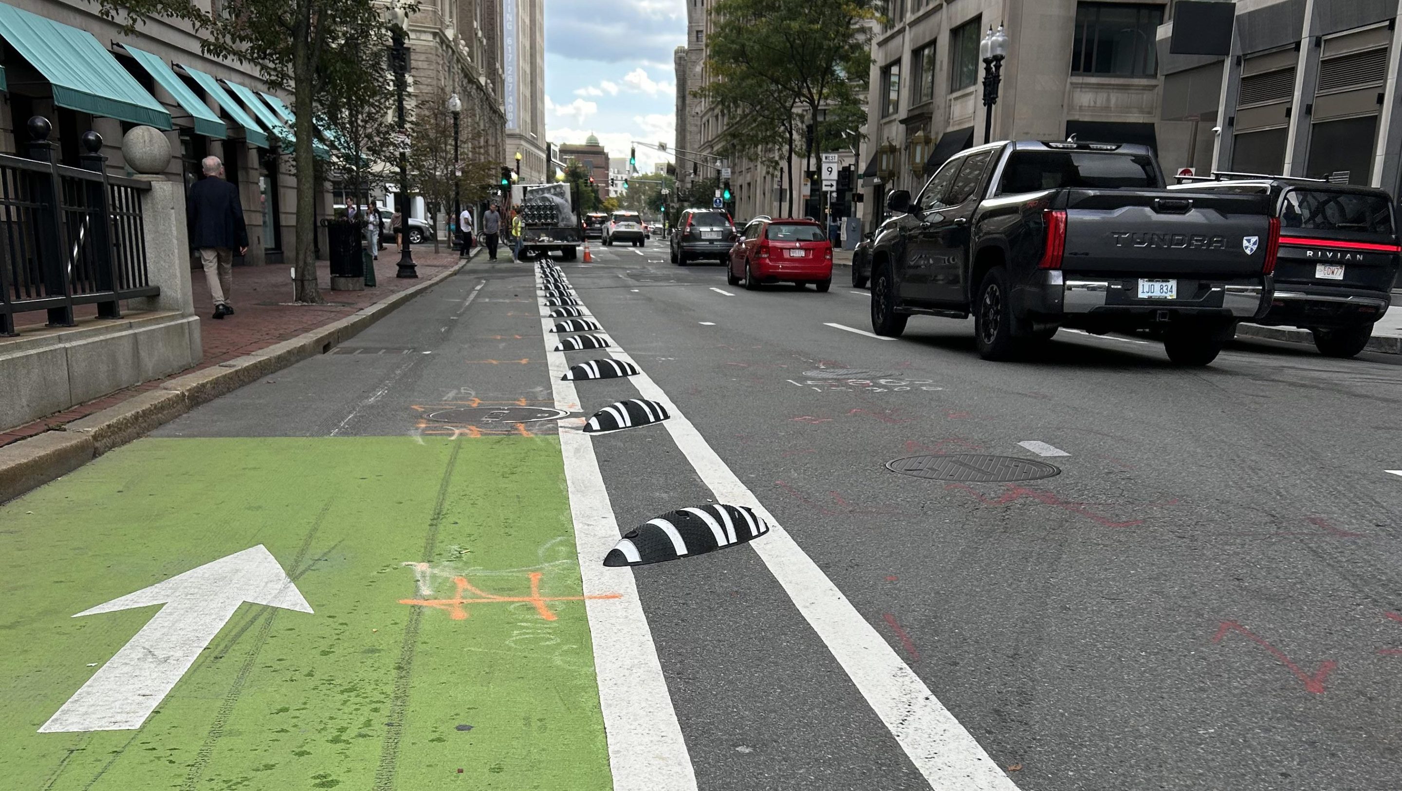 A low-angle view of a city street with a green-painted bike lane to the left, and a row of black-and-white striped plastic mounds installed between two white stripes in the buffer zone between the bike lane and the car lanes to the right. In the distance, workers are installing more of the black-and-white barriers from a truck parked in the bike lane.