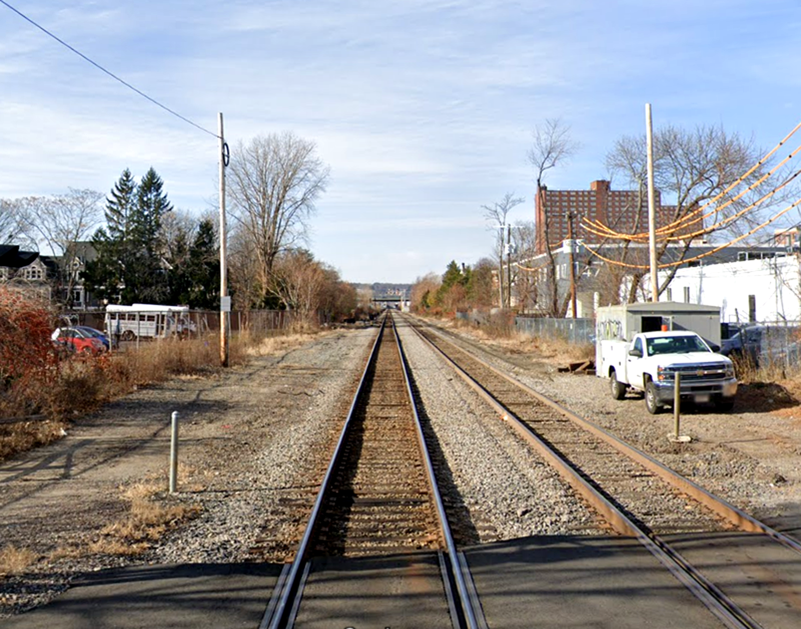 A look at the Fitchburg Commuter Rail Line from the Sherman Street grade crossing in Cambridge, Massachusetts. The train tracks with a white utility truck and industrial buildings on the right and trees, shrubs, and a small parking lot on the left.