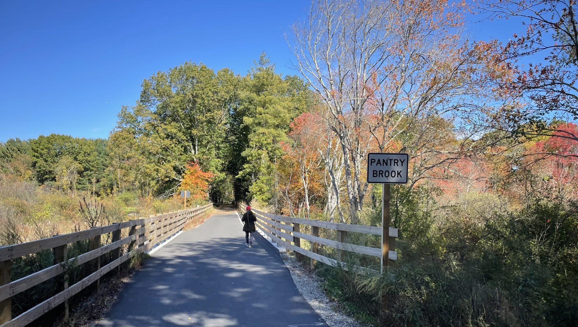 A woman in a black coat and orange hat walks over a short bridge on a straight paved trail lined with wooden fences. The trail runs through a meadow lined with trees with bright red and orange fall leaves. A sign next to the bridge reads "PANTRY BROOK"