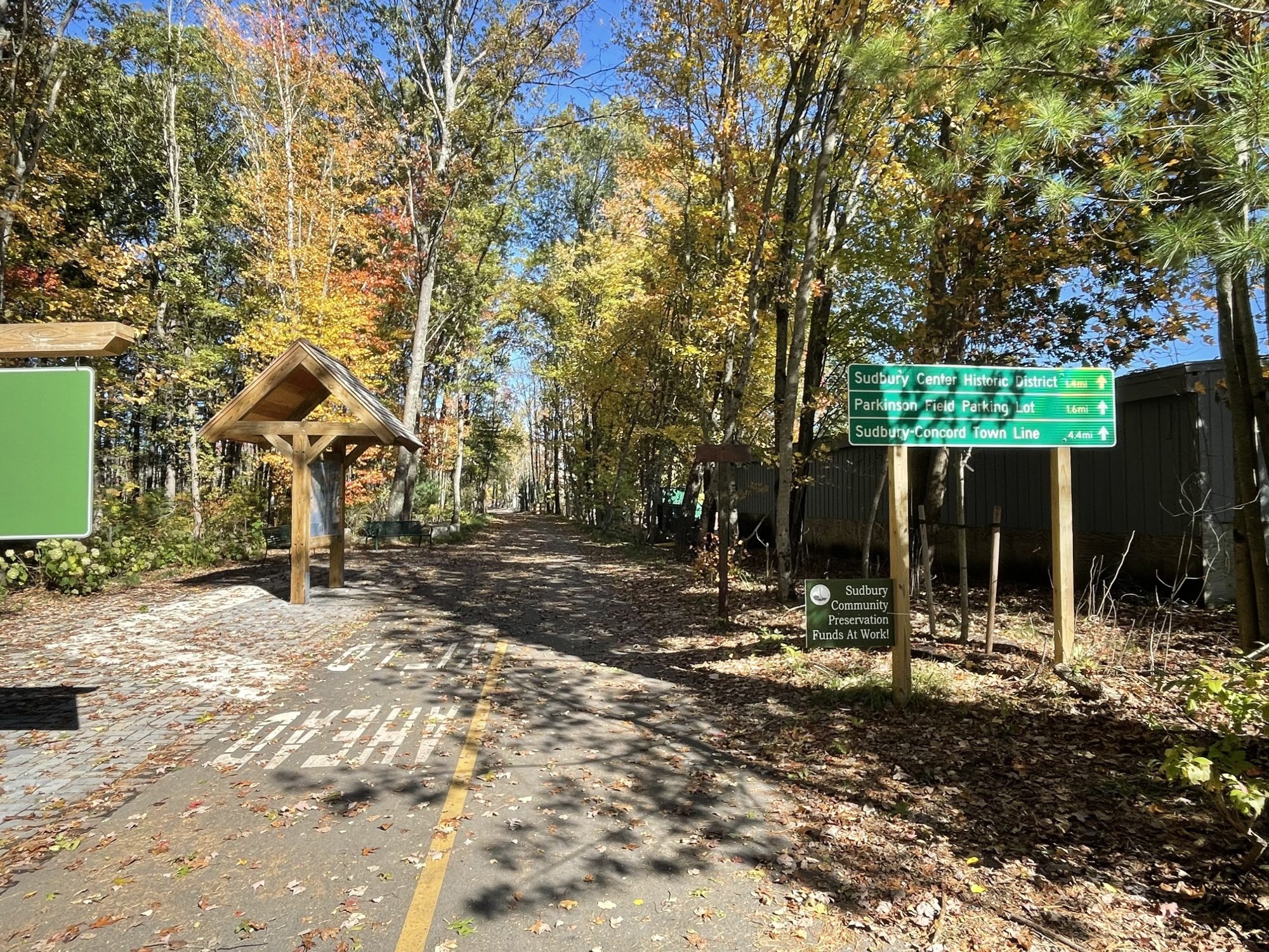 A long, straight bike trail cuts through the woods under fall leaves with a trail kiosk to the left and a green sign to the right that reads "Sudbury Center Historic District, 1.4 miles; Parkinson Field Parking Lot, 1.6 mi, Sudbury-Concord Town Line, 4.4 mi."