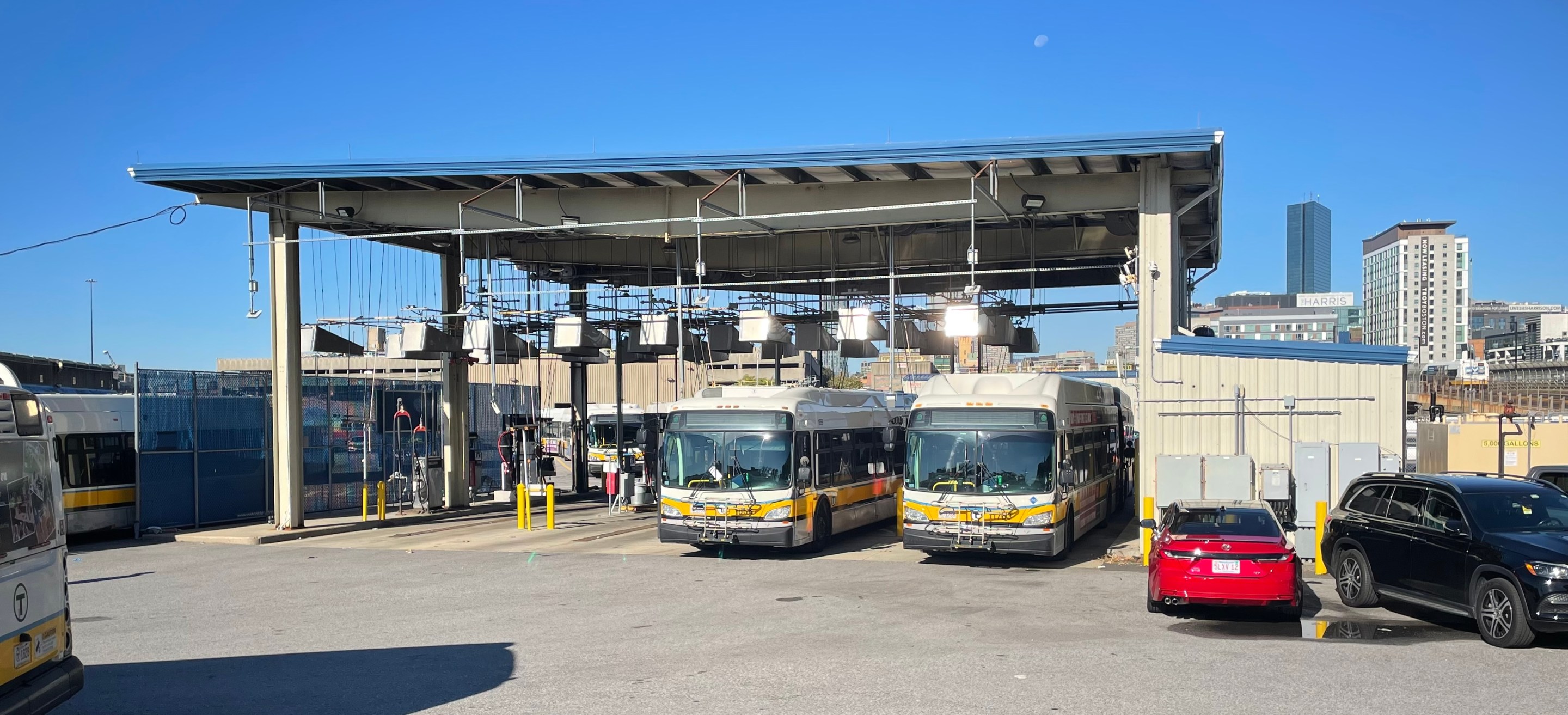 Two MBTA buses refueling under a large metal canopy under a blue sky