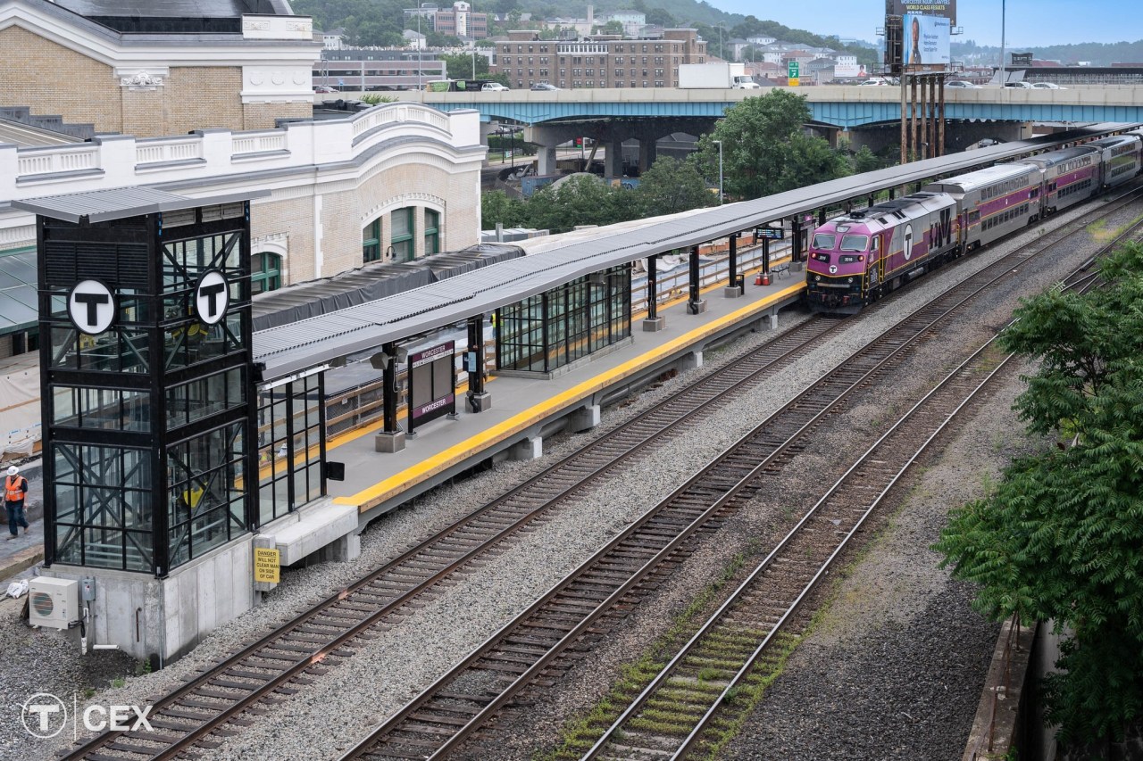 An elevated view of an MBTA commuter rail platform, with the T logo on the side of an elevator shaft at the near end of the platform in the foreground, and a purple MBTA commuter rail train pulling into the station in the distance at the top right edge of the image. To the left is the historic Worcester Union Station building and the I-290 highway viaduct runs along the top edge of the image.