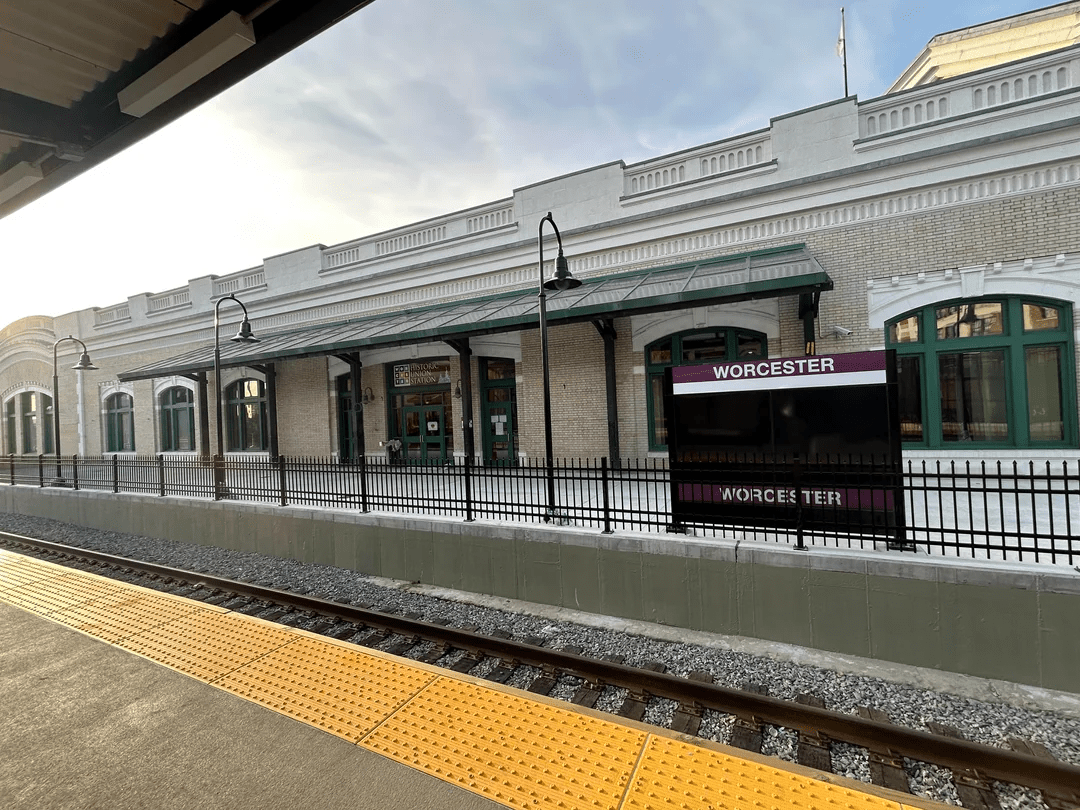 A photograph of a train platform in front of a historic station building. A platform in the foreground features a yellow tactile warning strip. The platform on the other side of the railroad track in blocked by a new fence. A sign directly behind the fence reads "WORCESTER" on a purple background.