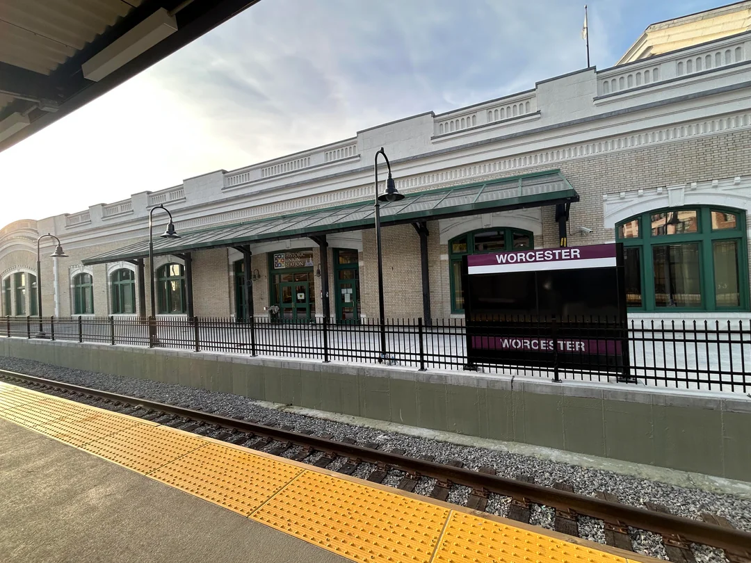 A photograph of a train platform in front of a historic station building. A platform in the foreground features a yellow tactile warning strip. The platform on the other side of the railroad track in blocked by a new fence. A sign directly behind the fence reads "WORCESTER" on a purple background.