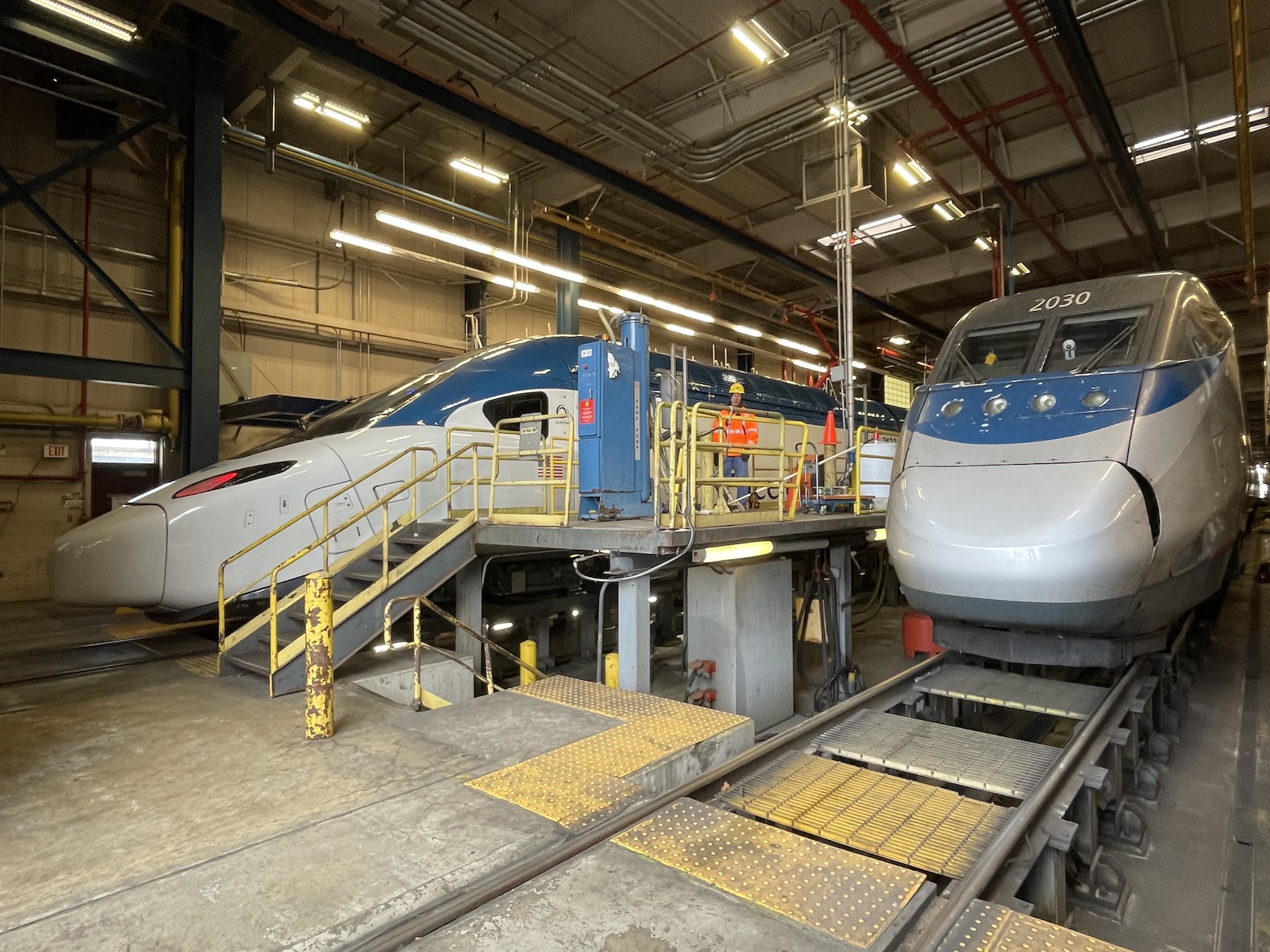 Two grey and blue trains with sloping noses are parked inside an indoor maintenance facility next to a high-level platform in the center.