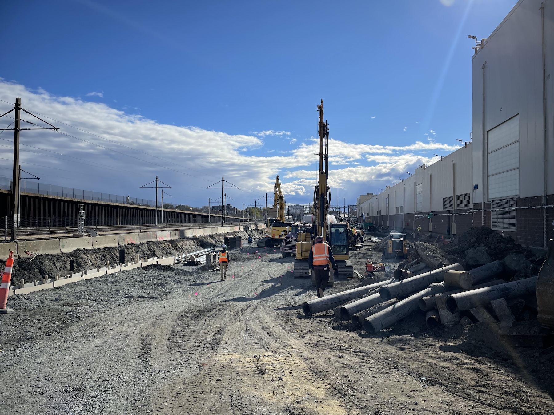 Large earthmoving equipment works in a dirt yard between a railway track (left) and a large, long warehouse building (right).