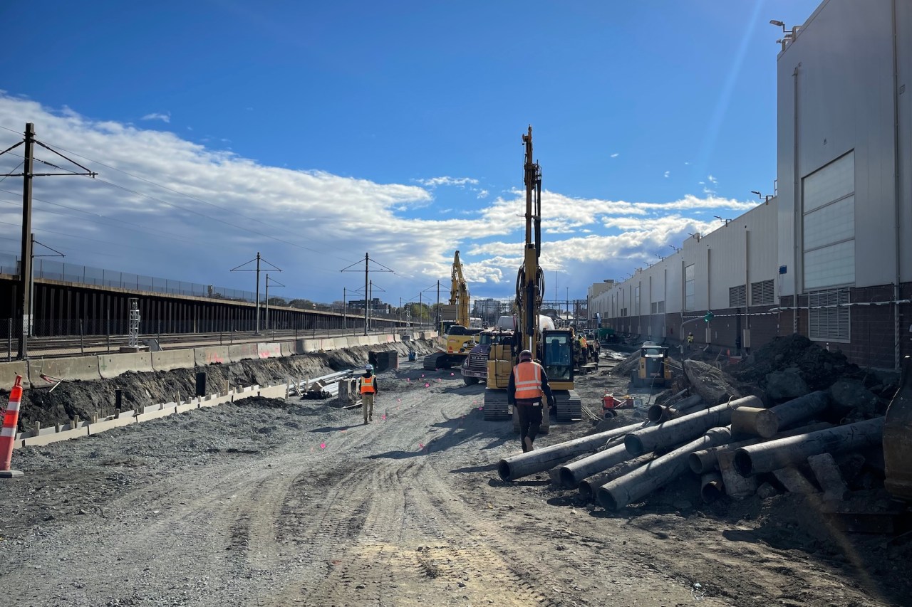 Large earthmoving equipment works in a dirt yard between a railway track (left) and a large, long warehouse building (right).
