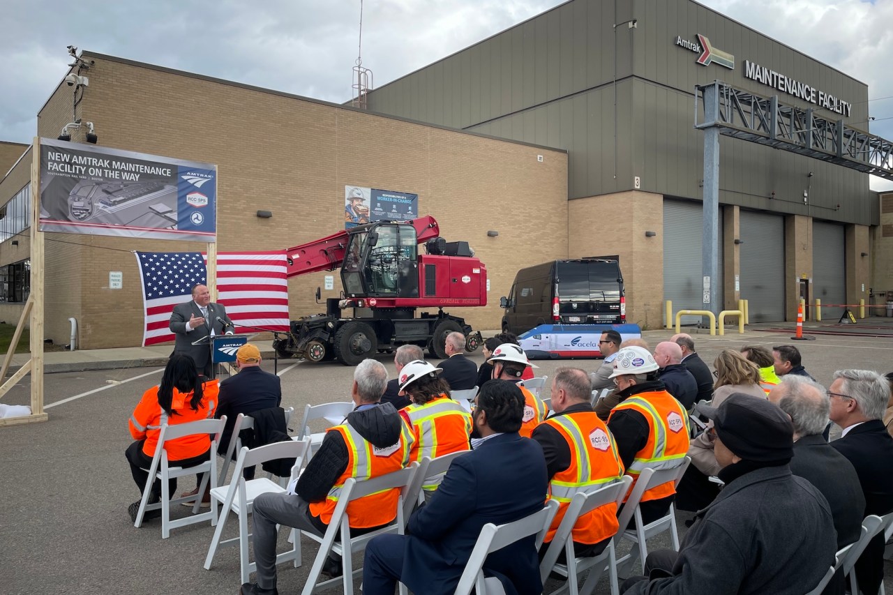 A small crowd seated in folding chairs, wearing overcoats and bright orange construction vests, faces a speaker at a podium in front of a large warehouse building with a sign that says "Amtrak Maintenance Facility." A banner behind the speaker says "New Amtrak Maintenance Facility On the Way". A model of the Acela train is in the background.