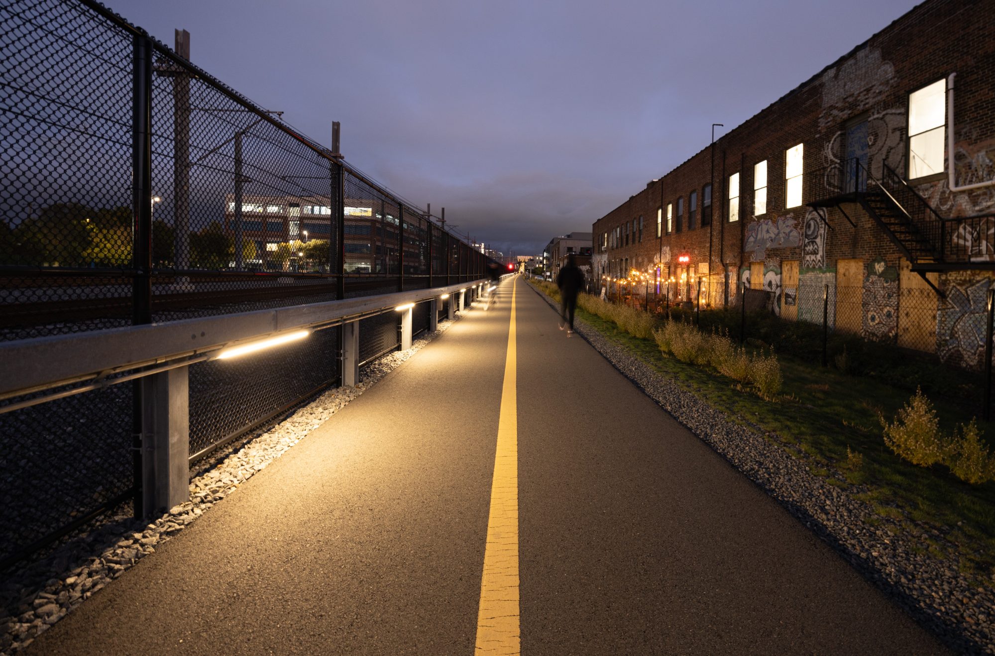 A view down a long, straight bike path at dusk. In the center is a painted yellow line stretching to the horizon; to the left is a chain link fence with bands of horizontal lighting illuminating the path at regular intervals from underneath a steel railing at waist height. On the right is a 2-story brick warehouse building with illuminated windows and graffiti art. In the middle distance are two people blurred from a long photo exposure walking along the path.