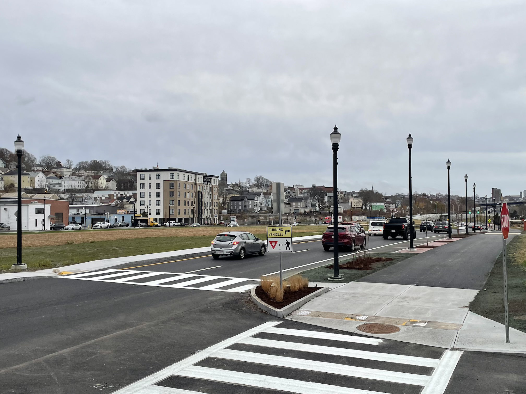 A view down a two-lane, one-way street with a wide shared-use path on the right side and a wide grassy lawn to the left. Across the lawn is another street lined with low-rise buildings, with one taller 6-story building standing out in the middle distance.
