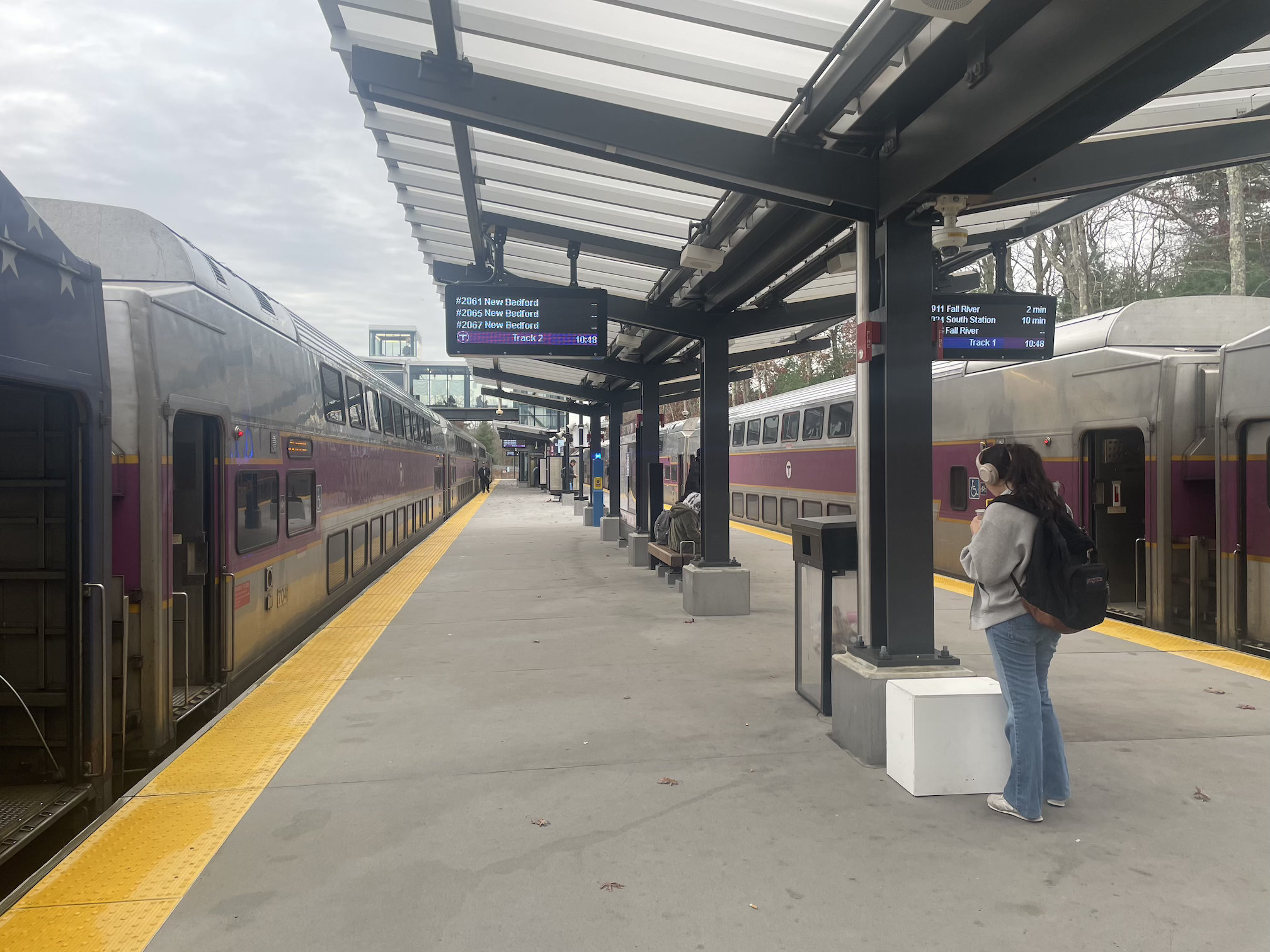 A handful of passengers wait on a train platform with a v-shaped roof canopy next to two waiting trains with the MBTA's purple stripe color scheme. Electronic signs above list upcoming departures for New Bedford and Fall River and South Station.