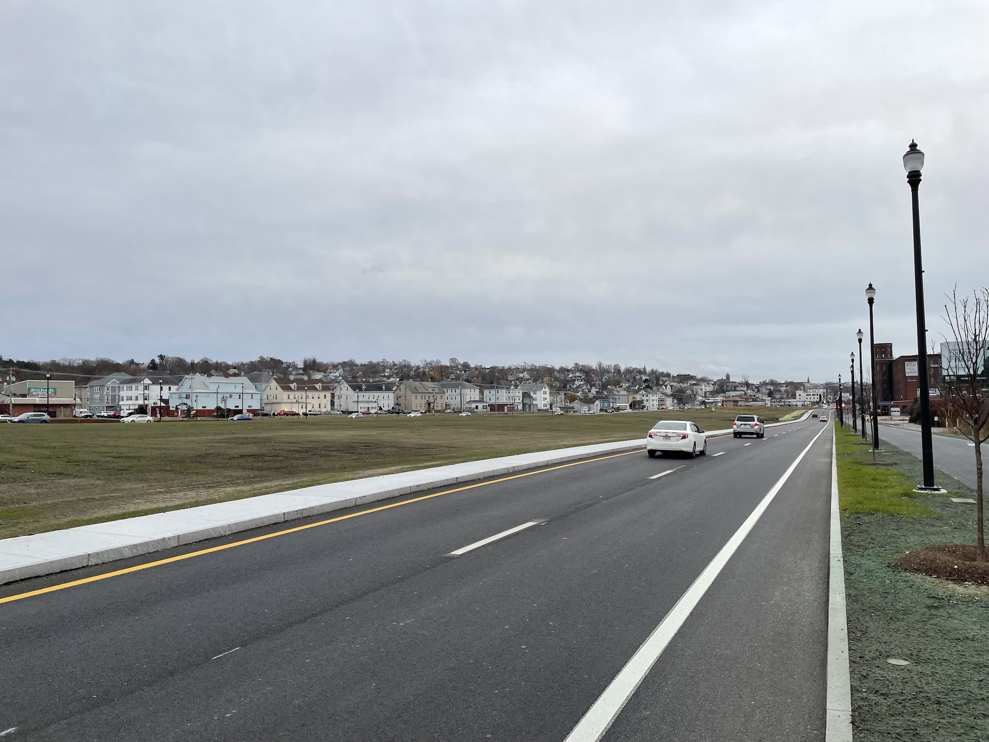 A view down a long, straight two-lane highway lined with street lamps and newly planted trees. To the left is a large grassy area, several hundred feet wide, with rows of multi-story houses in the distance on the other side.