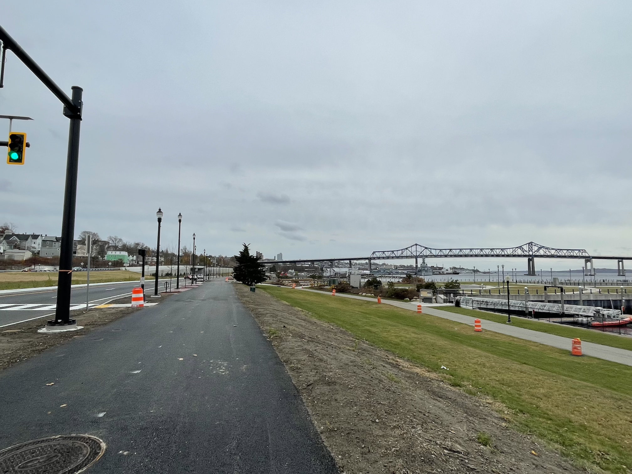 A view down a long, straight shared-use path. To the left is a two-lane roadway with a traffic light in the foreground. To the right is a slope that leads down to a dock on a waterfront area. In the distance is a tall highway bridge.