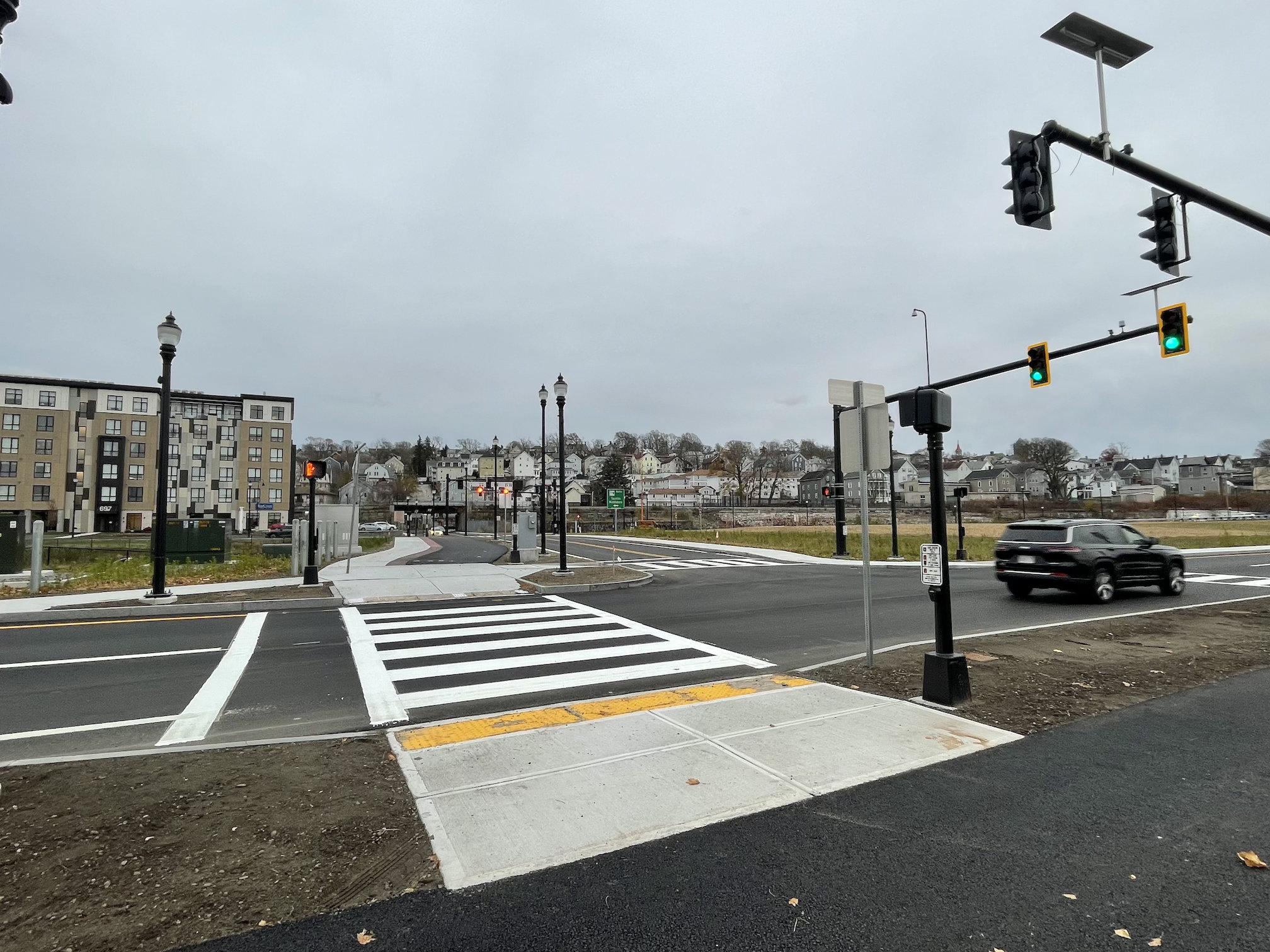 An intersection of two city streets with a broad crosswalk in the foreground and a pair of traffic lights suspended above the intersection at right. In the distance is visible a railroad overpass and beyond that a residential neighborhood of small 2- and 3-story apartment buildings on a hillside above the railway.