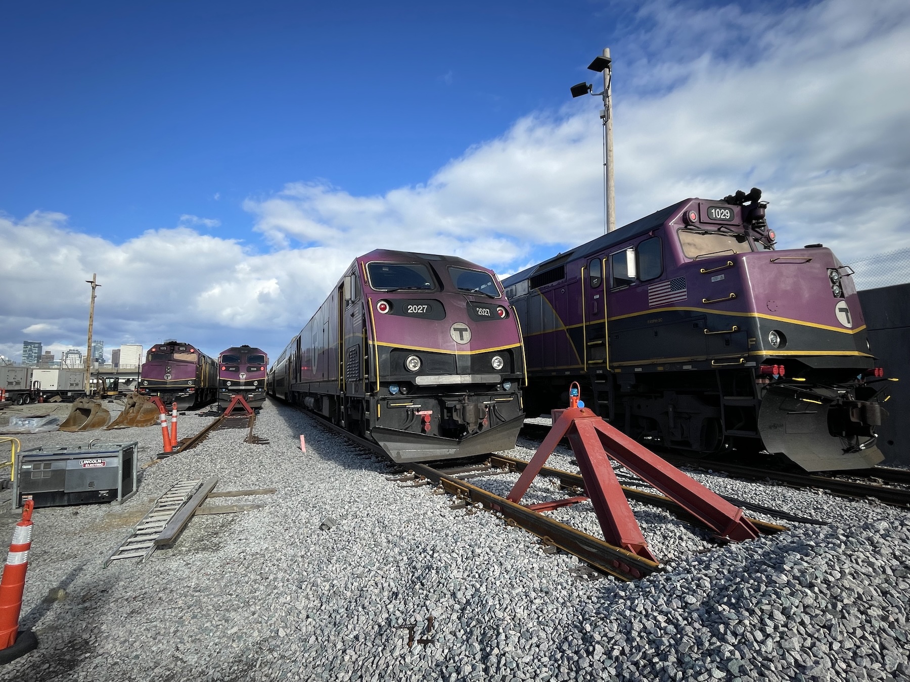 Four purple MBTA diesel locomotives parked next to each other on storage tracks in a large layover yard covered with ballast rocks. The Boston skyline is visible in the distance on the left edge of the photo.