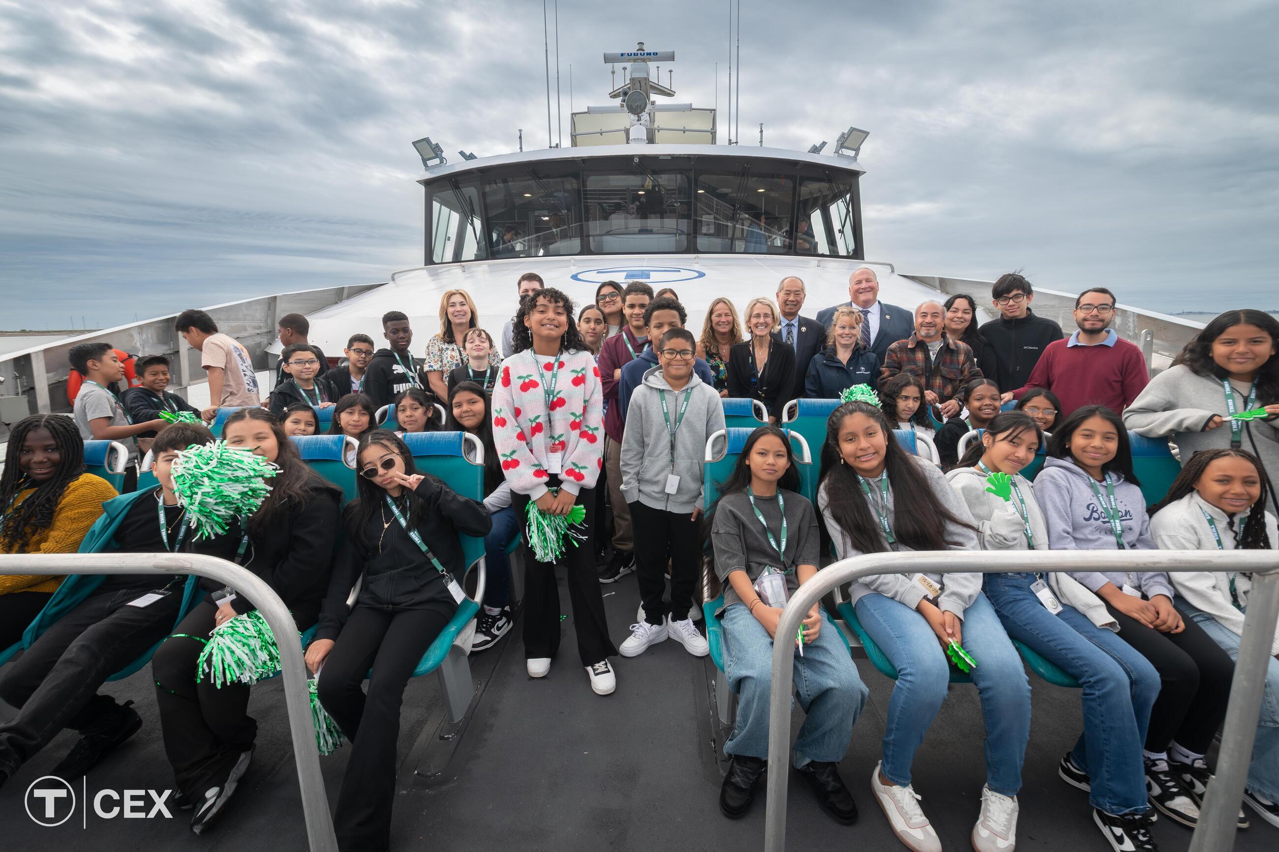 A group of students and adults pose for a photo on the deck of an MBTA ferry with cloudy skies overhead.