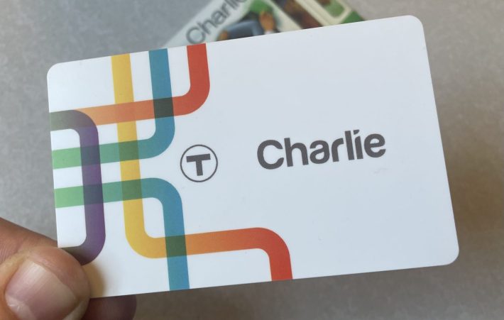 A plastic credit-card sized card, mostly white, with the T logo next to the word "Charlie" in a sans-serif font. On the left side of the card are several interwoven colored bands, evocative of the MBTA subway map.