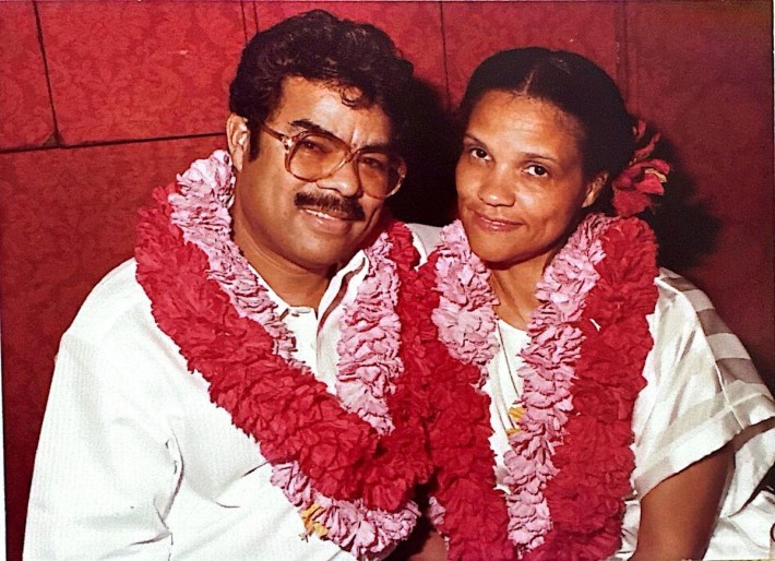 An older color photo shows a brown-skinned man and a woman grinning and leaning into each other with large floral leis around their necks. 