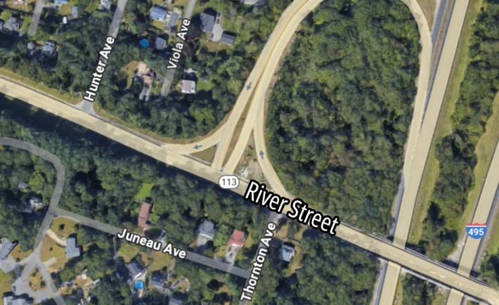 A satellite view of a highway interchange. In the center running from upper left to lower right is a straight road labelled "River Street". Smaller side streets branch off from River St. into suburban neighborhoods to the left.