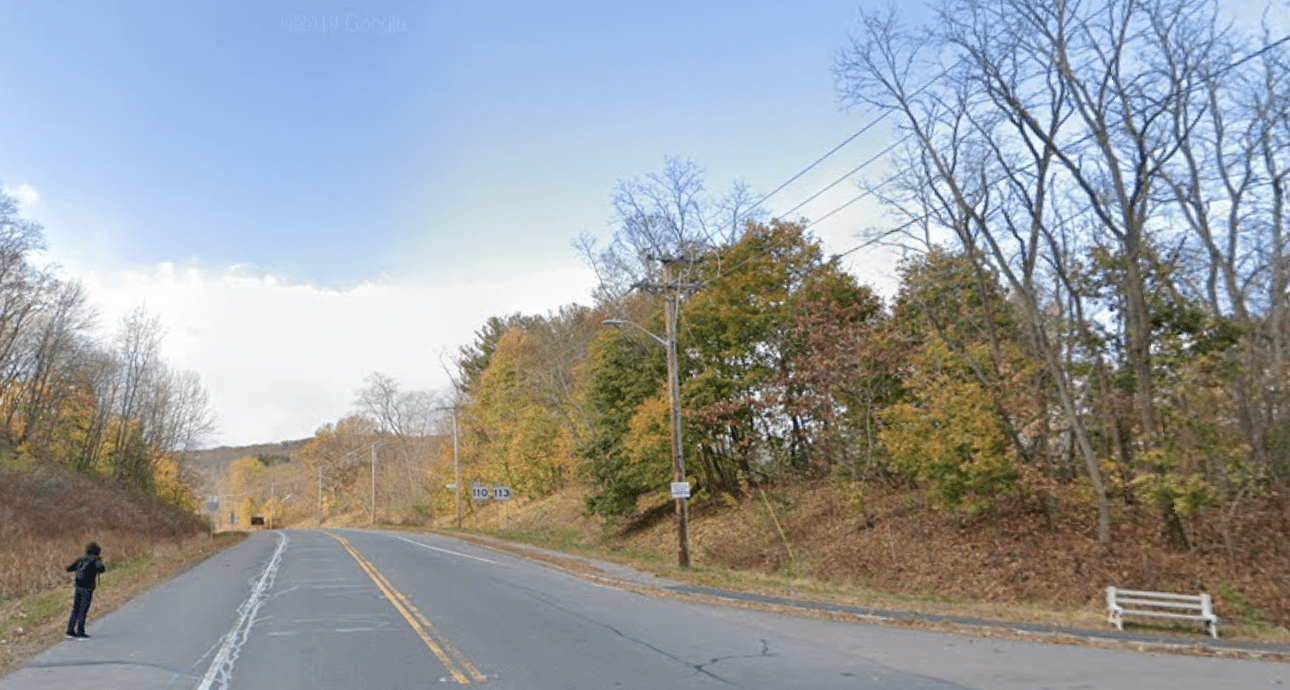 A view of a wide 2-lane highway through a wooded area in the autumn. On the left, a man stands on the roadside, where there is no sidewalk. On the right side of the road is a bench alongside a curb.