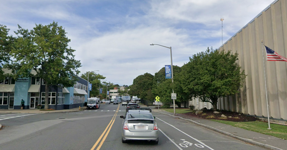 A view down a two-lane city street lined with on-street parking. A painted bike lane is on the right, and a "25 mph" speed limit sign. A large industrial building dominates the right side of the image, and on the left side of the street is a senior center.