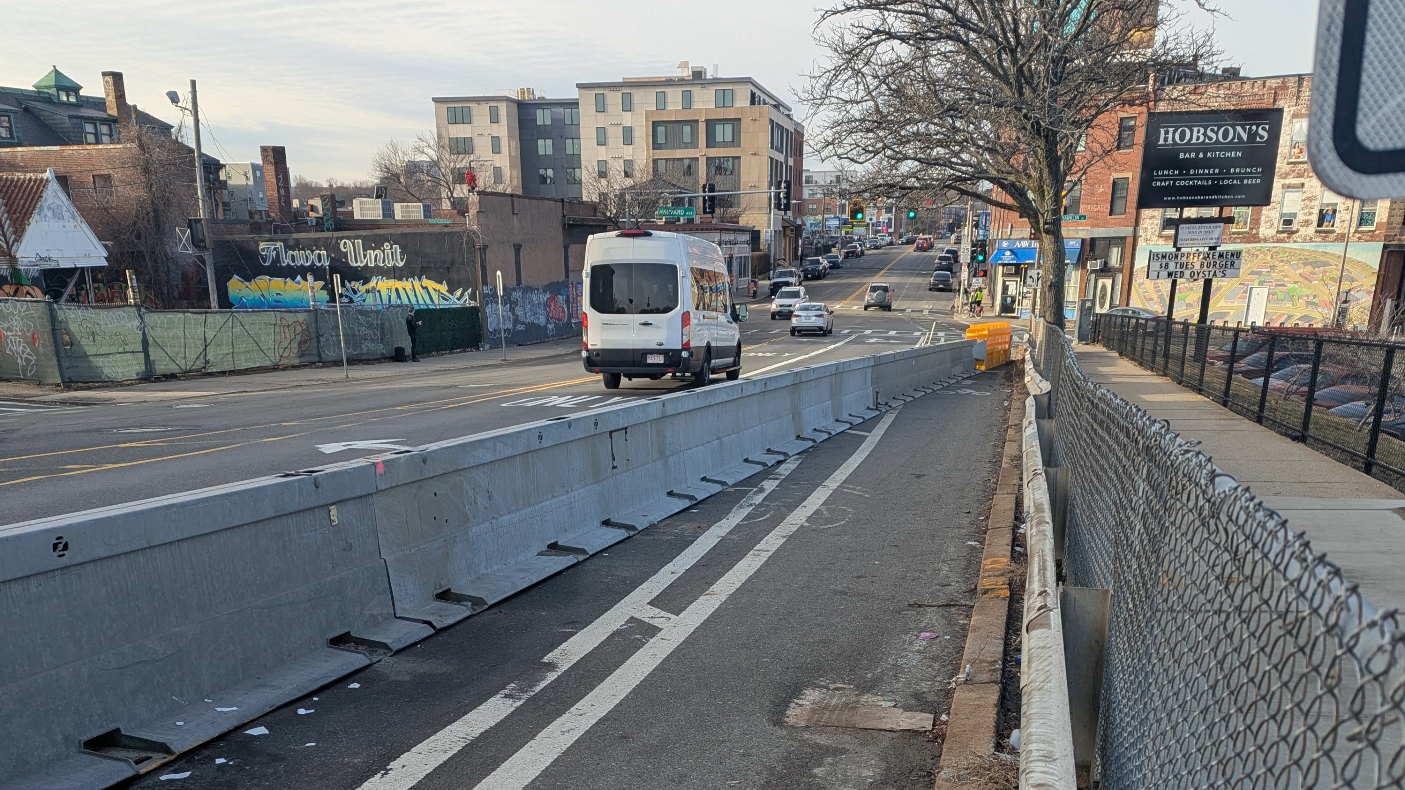 A bike lane on a wide city street in winter is blocked by a highway crash barrier that extends from the lower left corner of the image to the curb in the middle distance.