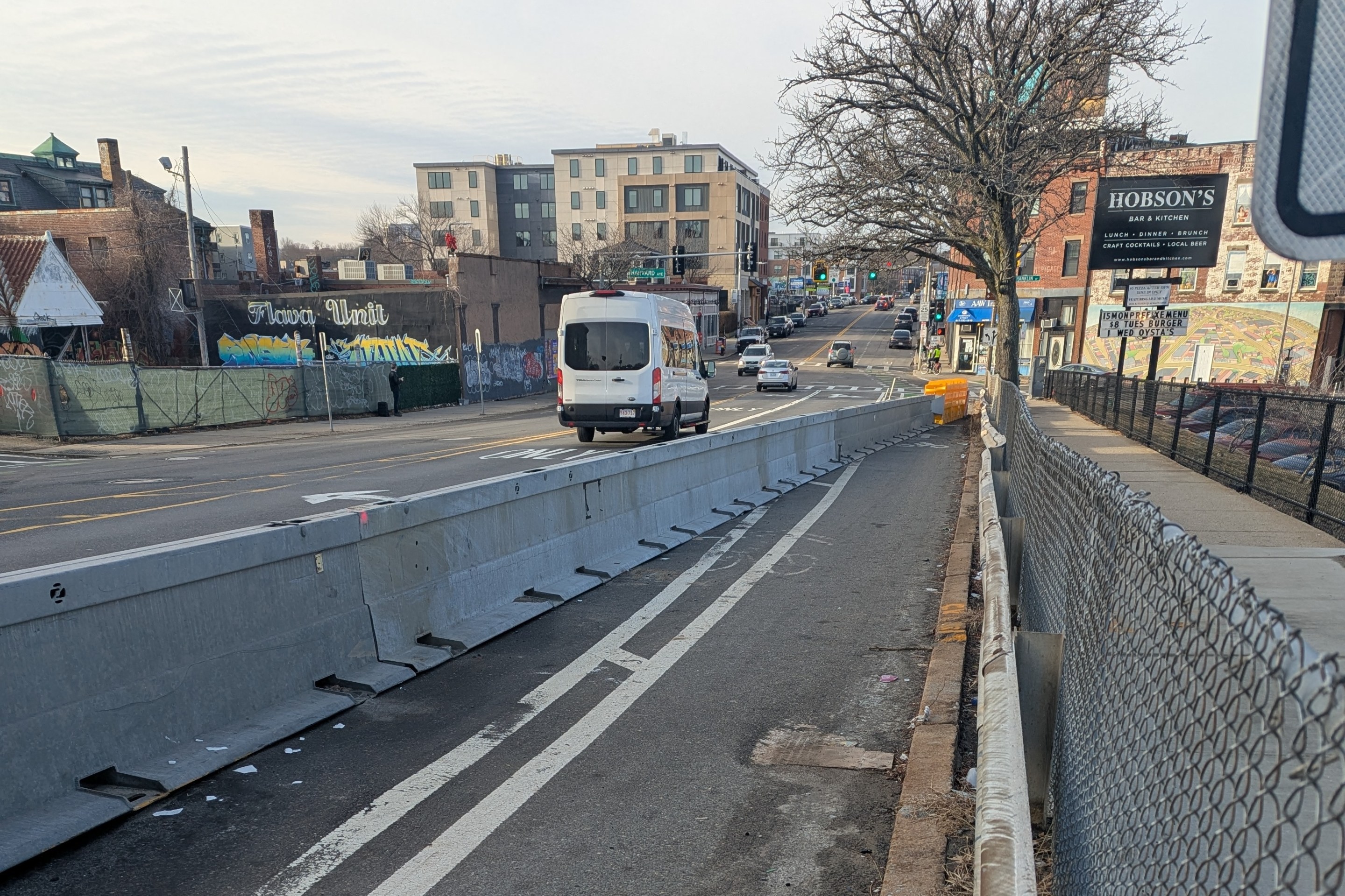 A bike lane on a wide city street in winter is blocked by a highway crash barrier that extends from the lower left corner of the image to the curb in the middle distance.