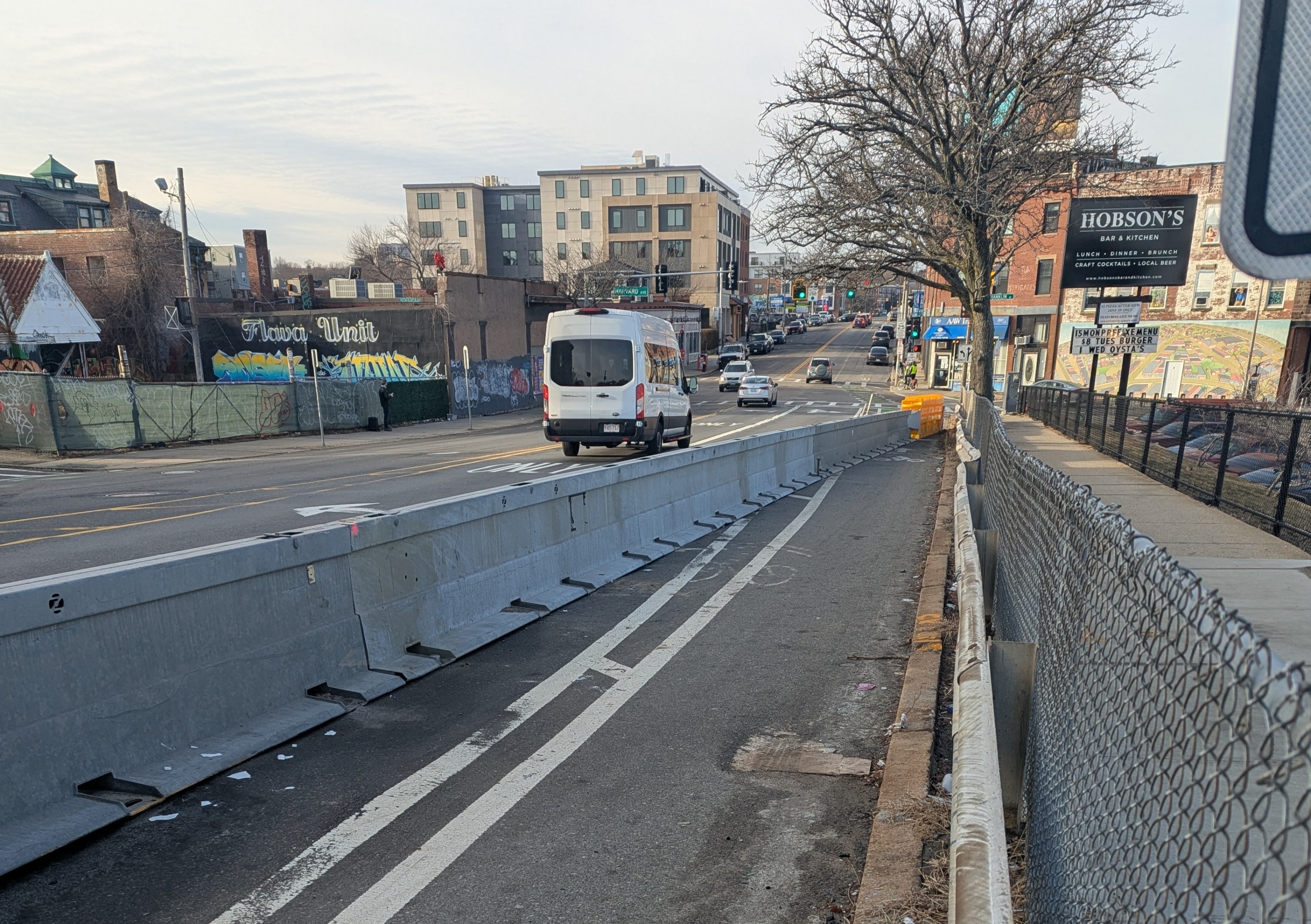 A bike lane on a wide city street in winter is blocked by a highway crash barrier that extends from the lower left corner of the image to the curb in the middle distance.