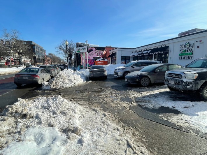 A messy snow-covered sidewalk blocked by cars parked in a parking lot next to a business with a sign that says "Baby Cafe"
