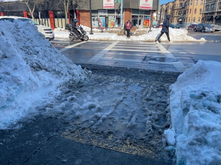 A low-angle photo of a crosswalk in winter, with the curb ramp in the foreground buried in several inches of slush and a median in the middle of the street blocked under a snowbank.