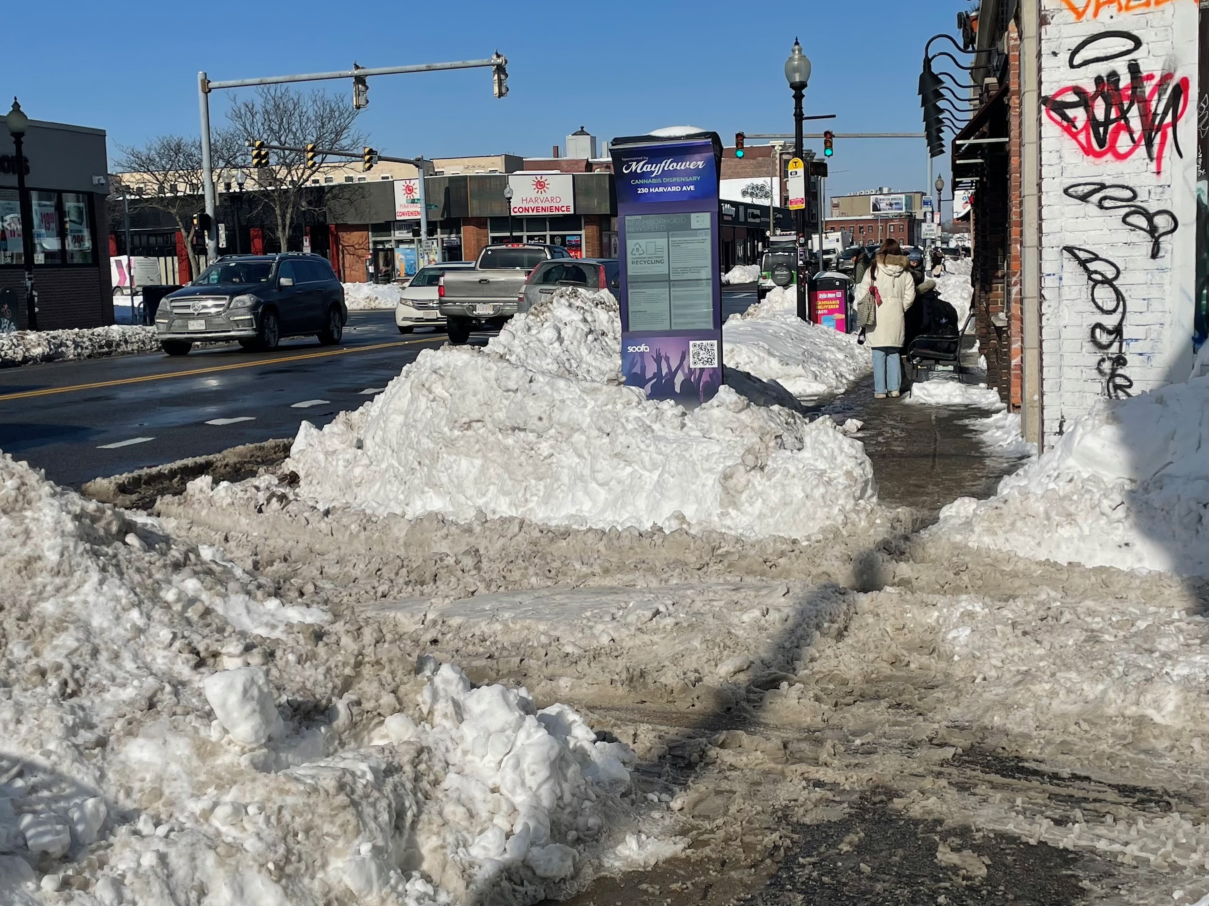 A sidewalk buried in a messy mixture of snow and slush along a busy city street in a neighborhood commercial area lined with one-story retail buildings.