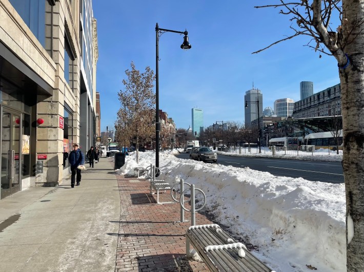 A broad city street in winter, lined with tall snowbanks, with a wide, well-cleared sidewalk on the left side of the image.