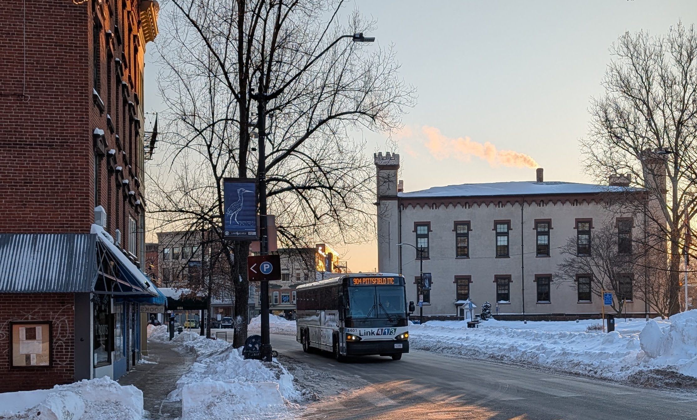 A coach bus with a headsign reading "904 Pittsfield TC" and a logo that says "Link413" drives down the main street of a small city on a sunny winter morning.