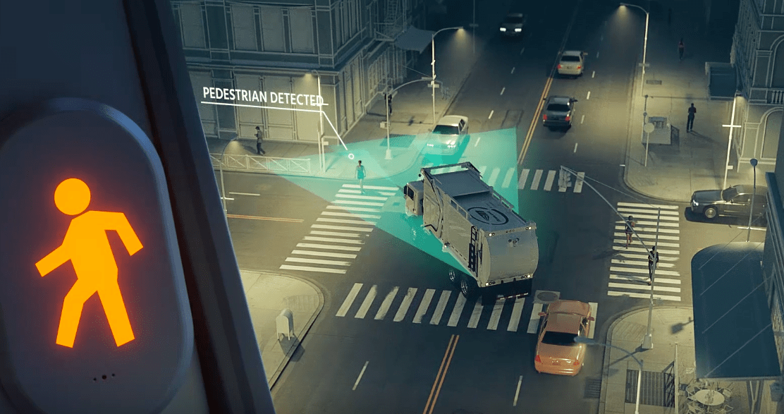 A computer animation shows a flashing pedestrian symbol next to an aerial view of a city street where a large dump truck is about to turn onto a side street where a pedestrian is crossing the crowwalk. A note above the crosswalk reads "pedestrian detected." A green triangle surrounding the truck indicates areas where cameras are detecting bikes and pedestrians in the truck's blind spots.