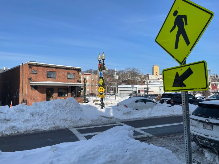 A city street lined with tall snowbanks, with a pedestrian crossing sign in the foreground at upper right, and a crosswalk that's blocked by snow.