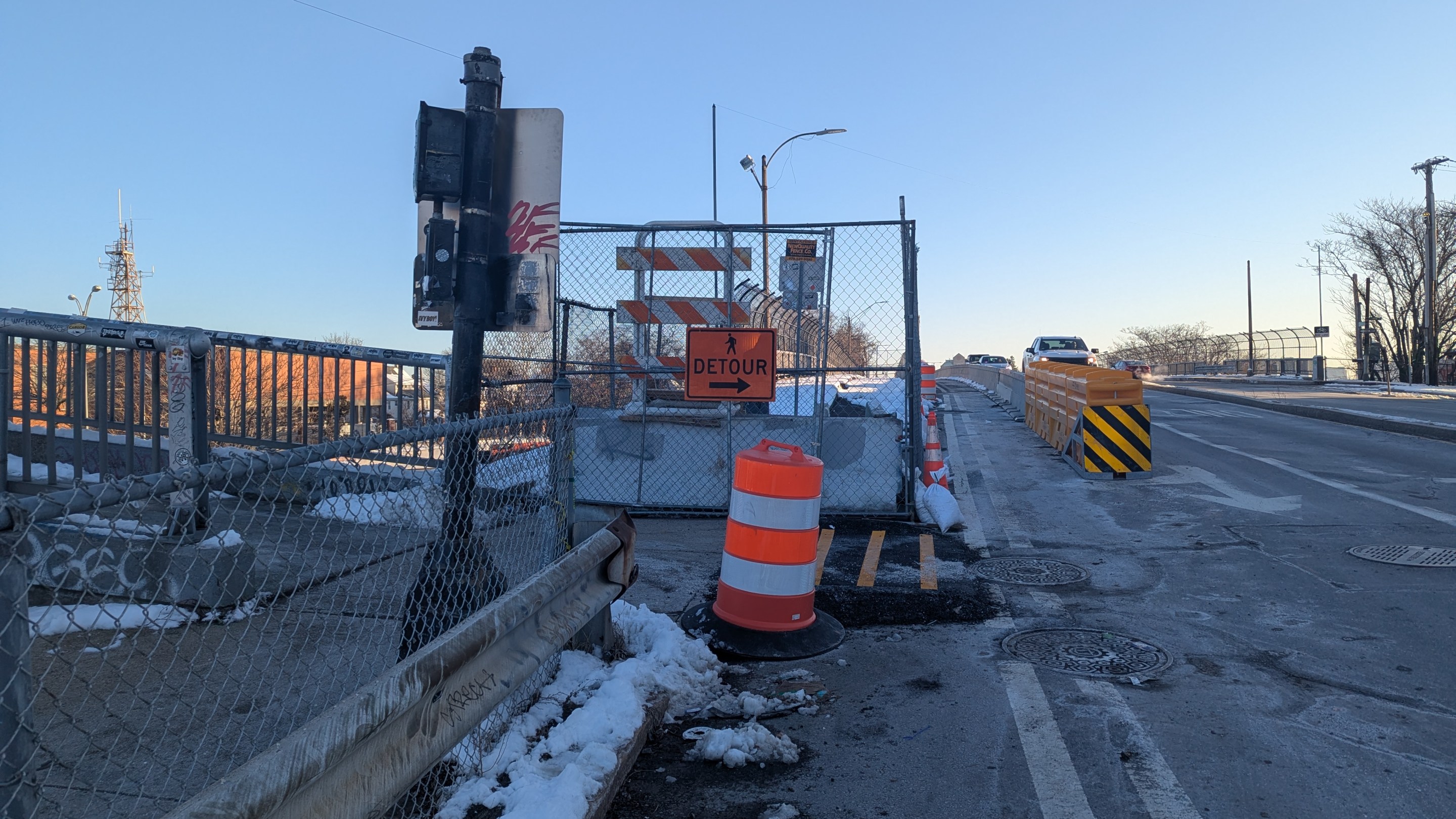A photograph of a bridge construction zone in winter. In the foreground a metal crash barrier and chain link fence separate a concrete sidewalk (left) from a multi-lane roadway (right). In the middle distance another fence blocks the sidewalk, with an orange "detour" sign pointing right towards the roadway behind an orange construction barrel. In the distance, a crash barrier in the roadway separates car traffic from a temporary sidewalk across the bridge. On the left edge of the photo a second pedestrian bridge joins with the sidewalk.