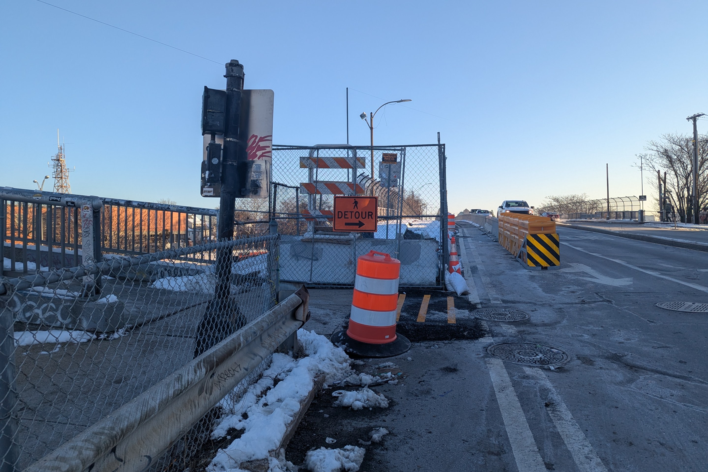 A photograph of a bridge construction zone in winter. In the foreground a metal crash barrier and chain link fence separate a concrete sidewalk (left) from a multi-lane roadway (right). In the middle distance another fence blocks the sidewalk, with an orange "detour" sign pointing right towards the roadway behind an orange construction barrel. In the distance, a crash barrier in the roadway separates car traffic from a temporary sidewalk across the bridge. On the left edge of the photo a second pedestrian bridge joins with the sidewalk.