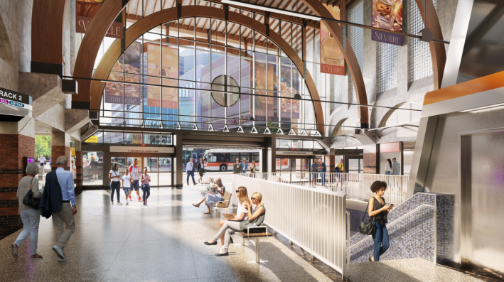 This rendering of Back Bay is from inside the station facing the street. A glass wall shows the Back Bay Logan Express bus passing by. Inside, there is a staircase with blue tiling that leads down to the train platforms. Commuters are walking up the stairs, through the station, and sitting down.