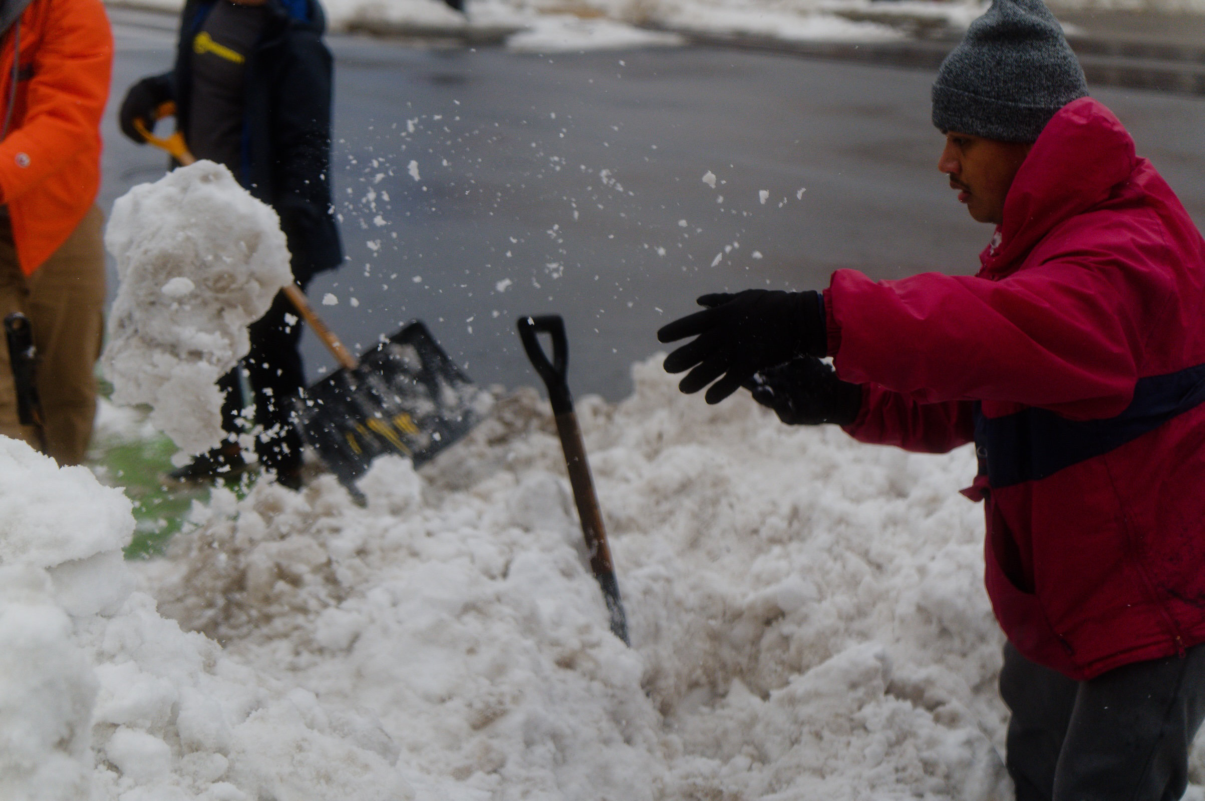 A brown-skinned man in a thick red winter parka and black hat tosses a huge chunk of snow aside as other volunteers behind him dig through a large snow bank.