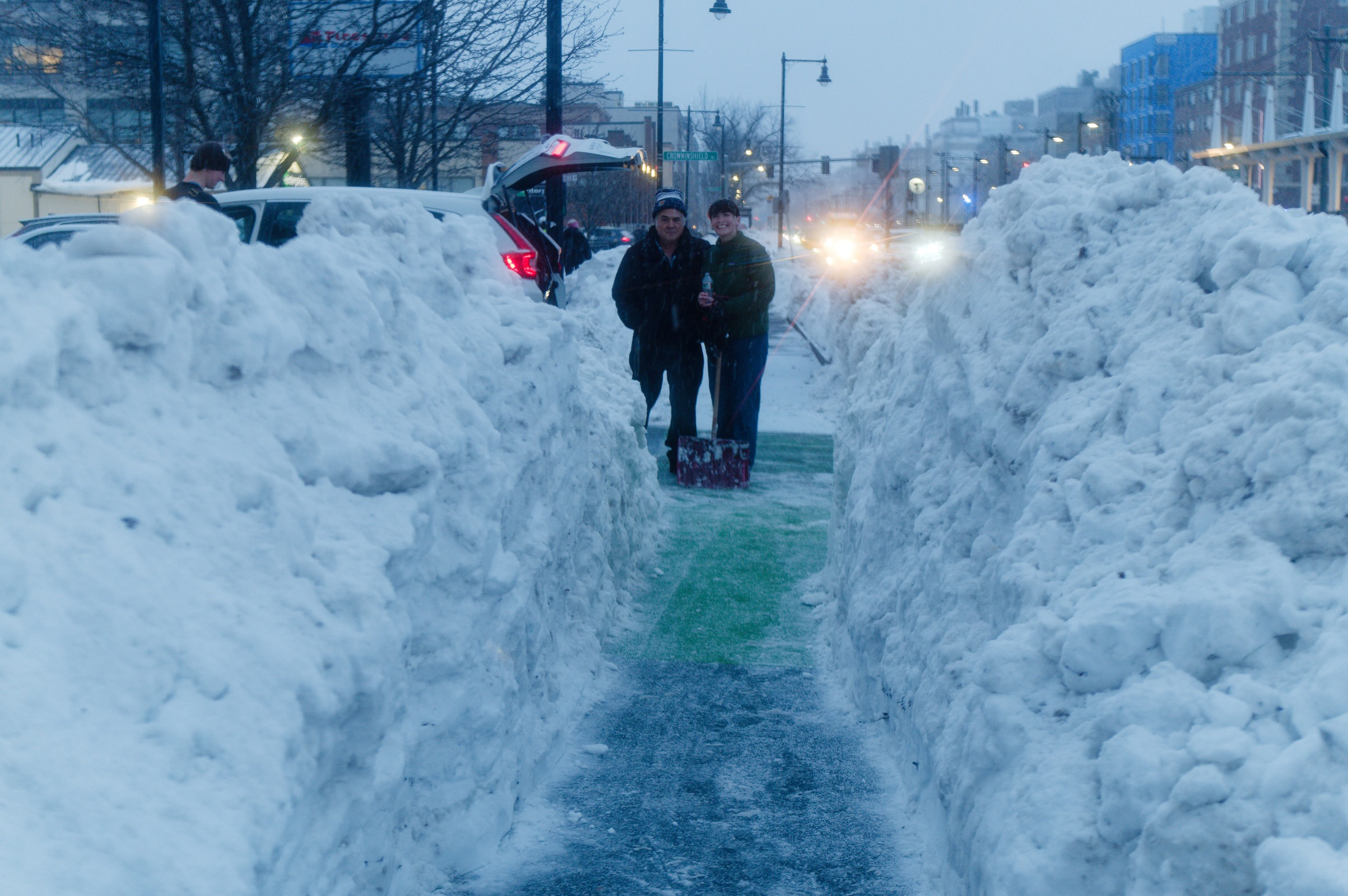 Two people with shovels stand at the end of a 5-foot-deep canyon of snow at the side of a wide city street on a winter evening