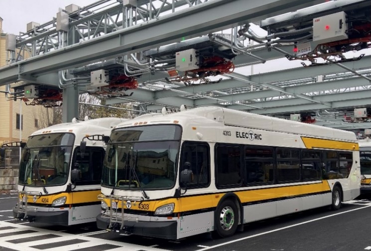 Two MBTA buses, white with yellow and black stripes under the windows and the word "ELECTRIC" above, sit in a parking lot under an elaborate steel gantry with charging equipment.
