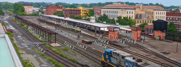 A view of a multi-track train station from an elevated perspective from a nearby building. Annotations overlaid on the image label each track, from Track 3 at left to track 8 at far right, with a CSX freight train approaching on Track 1 from the lower right. The abandoned Platform A is labelled at left, proceeding alphabetically to Platform D at upper right. In the distance are multi-story masonry buildings of a small city.