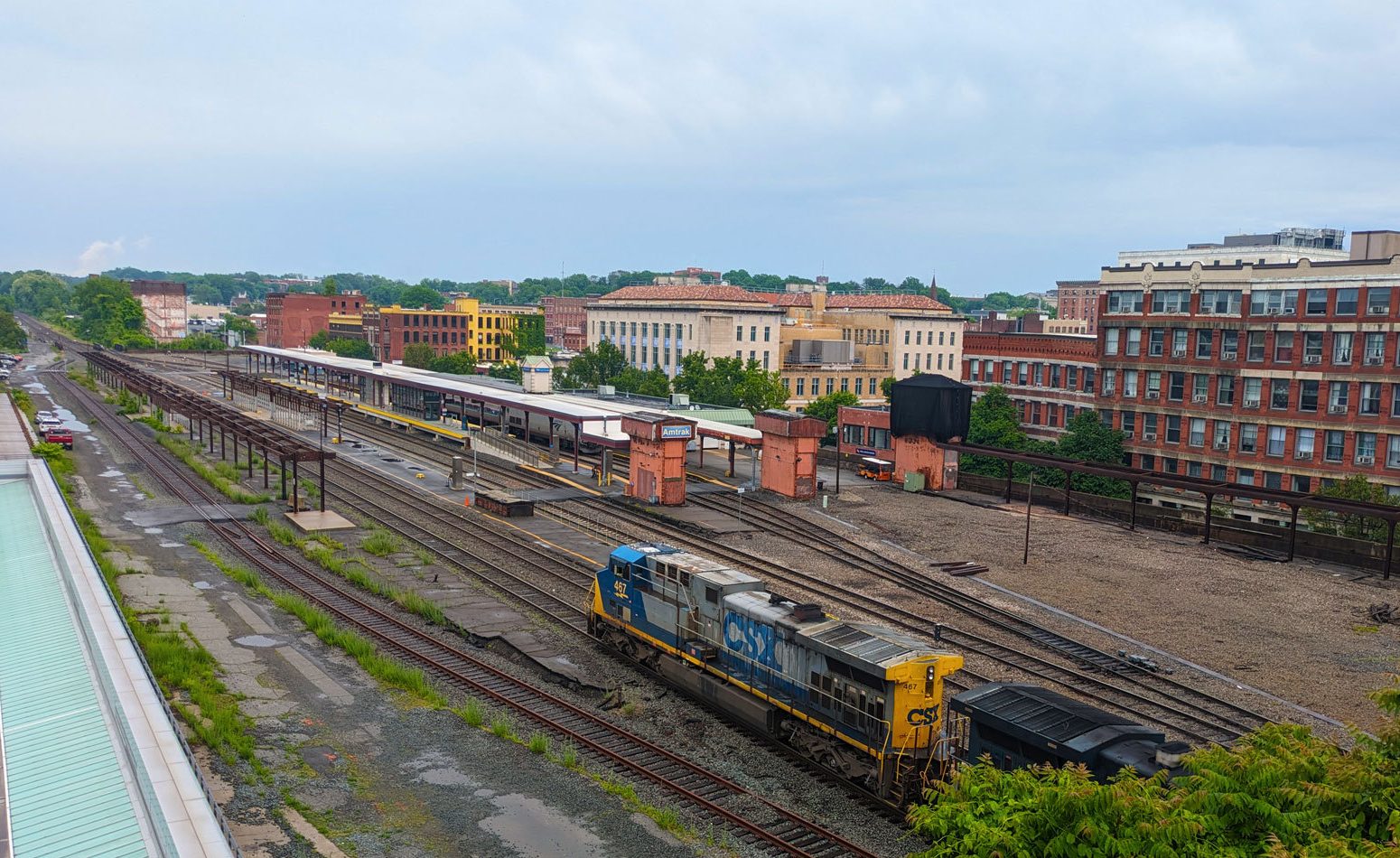 A view of a multi-track train station from an elevated perspective from a nearby building. A CSX freight train is approaching the platforms from the lower right. Several of the platforms at left are abandoned but there is a newer platform visible in the upper part of the rail yard. In the distance are multi-story masonry buildings of a small city.