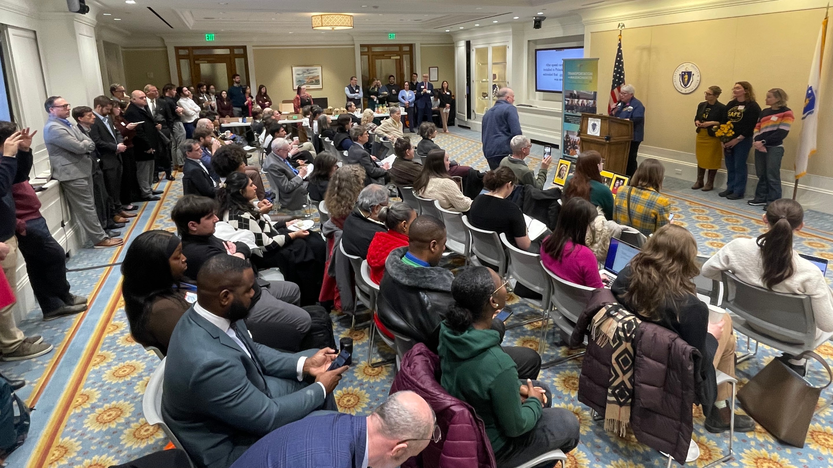 A crowd of approx. 60 people, some seated in four rows of chairs, and more standing against the walls, fills a room facing a podium where a white-haired woman is speaking in front of the Massachusetts state seal and two flags.