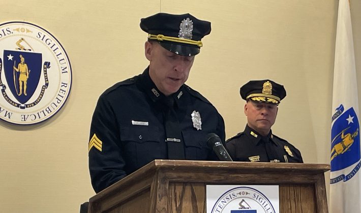 Two cops in uniform speak at a podium with the state seal of Massachusetts