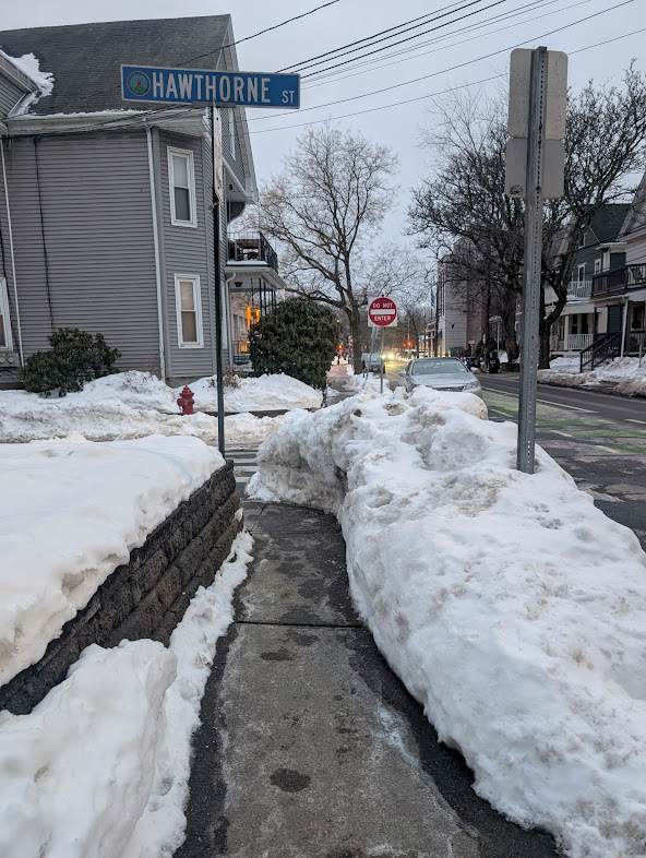Snow-lined sidewalk at Hawthorne Street in Porter Square, with tall snowbanks, a blue Hawthorne St street sign, and residential houses.